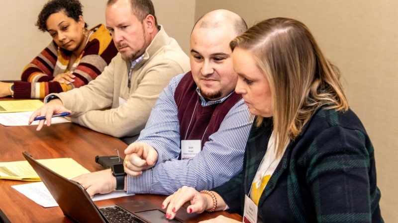 Four people sit at a wooden table. One man points at a laptop screen.