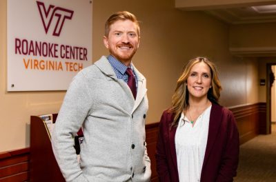 Matthew Fullen and Joanna Collins pose for a photo in a hallway with the Virginia Tech Roanoke Center logo on the wall behind them.