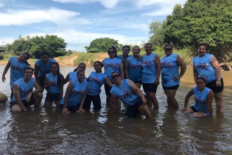 Group of 13 women and one man wearing matching Zumba t-shirts posing for a picture in a  small body of water.