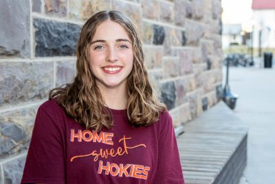 Kirsten Perrin wears a maroon shirt that reads "Home Sweet Hokies" as she sits for a portrait in front of a Hokie Stone wall. 