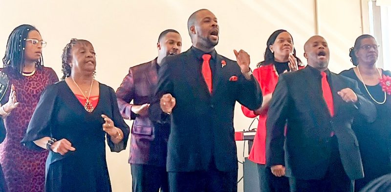 A group of people wearing black and red sing and dance in a line at the front of a room.