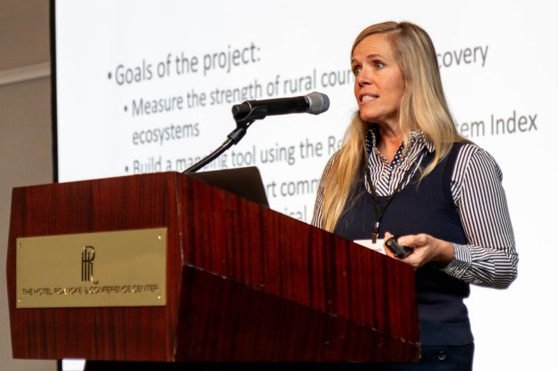 A woman stands at a podium with a presentation slide projected on a screen behind her.